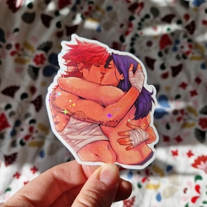 May include: A sticker featuring a colourful illustration of two women kissing. The women are drawn in a cartoon style with red and purple hair. The sticker has a glittery finish.