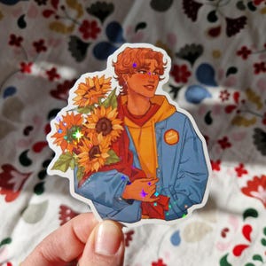 May include: A sticker featuring a cartoon illustration of a person with brown hair holding a bouquet of sunflowers. The person is wearing a blue denim jacket over a yellow hoodie. The sticker has a holographic finish.
