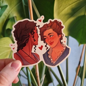 Puede incluir: Dos mujeres con cabello castaño se representan en una ilustración de primer plano. La mujer de la izquierda tiene el cabello castaño oscuro y lleva un top sin mangas azul oscuro. La mujer de la derecha tiene el cabello castaño claro y lleva una camisa azul y blanca con volantes. Ambas mujeres sonríen y se miran. Hay pequeños corazones rojos dispersos alrededor de las mujeres.