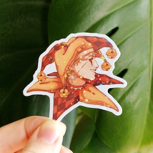 May include: A sticker featuring a stylized illustration of a jester. The jester wears a red and orange diamond-patterned hat with bells. The jester's face is detailed with makeup and a star design. The sticker has a holographic finish.
