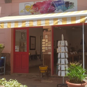 Pode incluir: Uma loja com uma marquise listrada de amarelo e branco. A loja se chama "Baz Art" e vende lembran&ccedil;as, presentes e artigos feitos &agrave; m&atilde;o. A placa &eacute; pintada em t&aacute;buas de madeira.