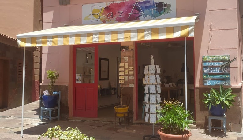 Pode incluir: Uma loja com uma marquise listrada de amarelo e branco. A loja se chama "Baz Art" e vende lembran&ccedil;as, presentes e artigos feitos &agrave; m&atilde;o. A placa &eacute; pintada em t&aacute;buas de madeira.
