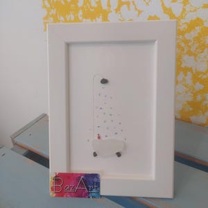 以下が含まれることがあります： 白い額縁に入った絵画。白い浴槽はシーグラスで作られており、青いシーグラスと白いシーグラスは水を、赤いシーグラスは浴槽にいる人を表しています。絵画は青と白の表面に置かれており、手前にBazArtのビジネスカードがあります。