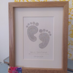 Op de afbeelding: Een ingelijst afbeelding met twee babyvoetjes. De afbeelding bevat de naam "RAUL SANTANA" en de datum "09.03.2024".