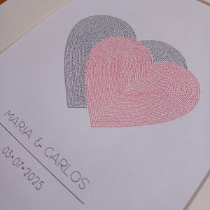 Op de afbeelding: Een ingelijste print met twee overlappende hartvormen bestaande uit herhaalde tekst. Het voorste hart is rood en het achterste hart is zwart. Onder de harten staat de tekst MARIA & CARLOS en de datum 03-07-2025 in zwarte inkt.