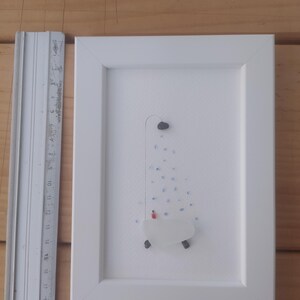 以下が含まれることがあります： シャワーヘッドと水滴がついたバスタブを描いた、シーグラスと石で作られた額装されたアート作品。作品は白い背景に描かれており、白いフレームで額装されています。