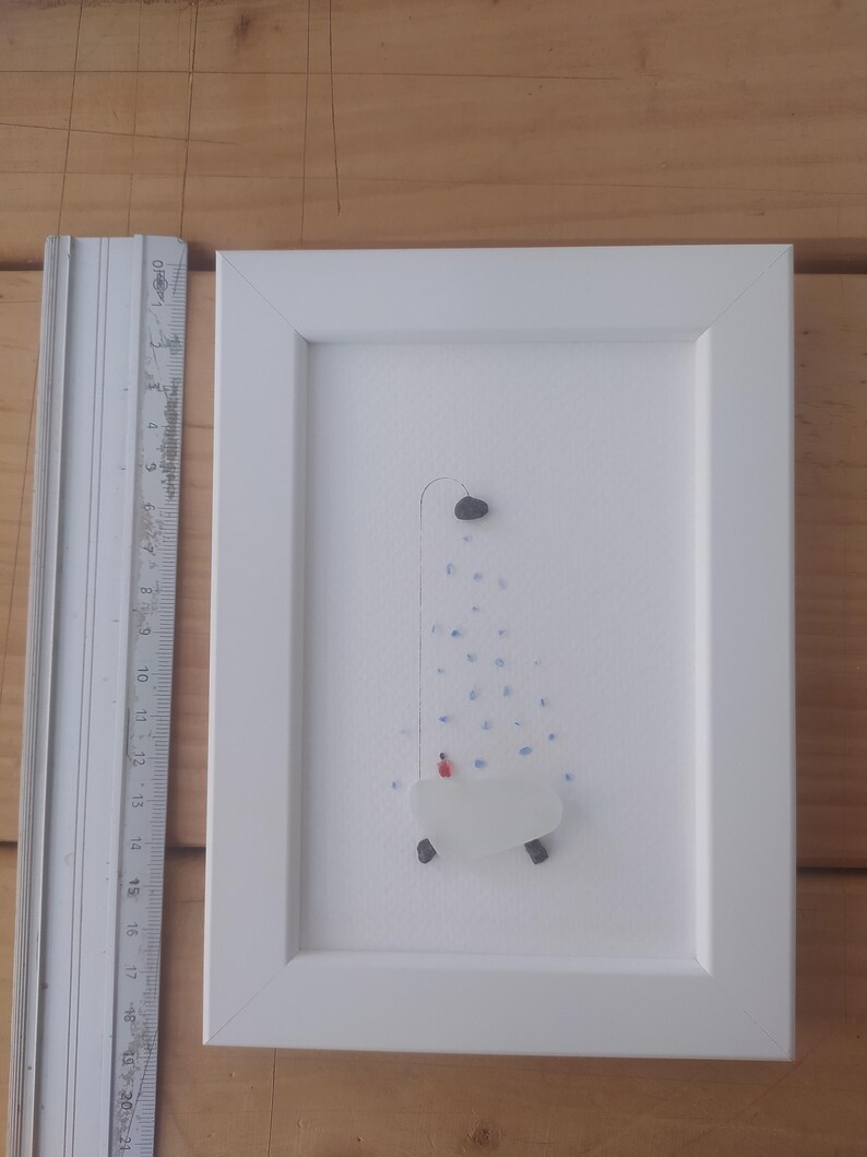 以下が含まれることがあります： シャワーヘッドと水滴がついたバスタブを描いた、シーグラスと石で作られた額装されたアート作品。作品は白い背景に描かれており、白いフレームで額装されています。