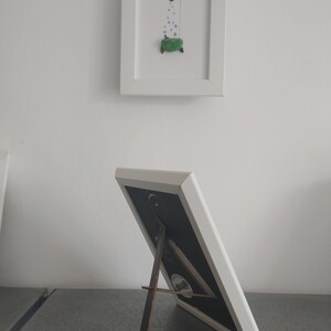 Backside of a white frame, standing on a table. The backside is black. The frame can be used standing or hanging on the wall.