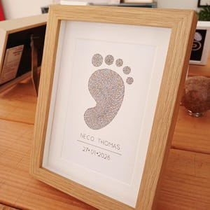 Ingelijst voorbeeld van een babyvoetje, gemaakt met handgeschreven woorden. Er onder de volledige naam van de baby en de geboortedatum.