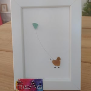 Puede incluir: Una imagen enmarcada en blanco con una obra de arte de vidrio de mar que representa un cochecito de bebé con una cometa atada a él. La obra de arte está sobre un fondo blanco. El marco tiene una etiqueta de BazArt en la parte inferior.
