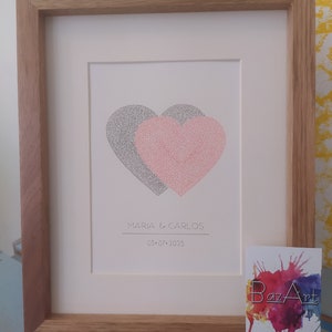 Op de afbeelding: Een ingelijst afbeelding met twee handgeschreven overlappende harten, een zwart en een rode, met de tekst "MARIA & CARLOS 03-07-2025" eronder. De afbeelding is in een houten lijst ingelijst.