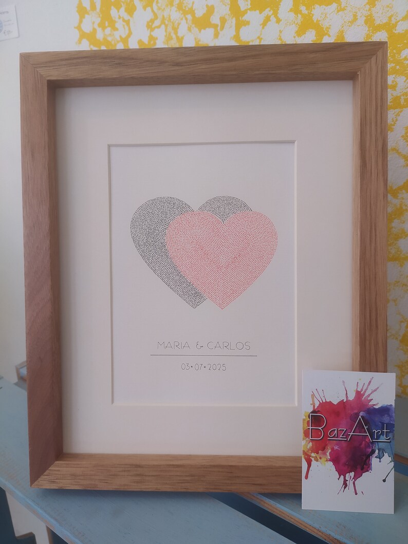 Op de afbeelding: Een ingelijst afbeelding met twee handgeschreven overlappende harten, een zwart en een rode, met de tekst "MARIA & CARLOS 03-07-2025" eronder. De afbeelding is in een houten lijst ingelijst.