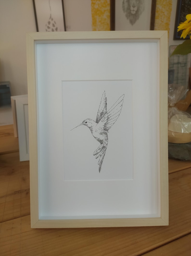 May include: A framed black and white line drawing of a hummingbird in flight. The drawing is on white paper and is framed in a light wood frame.