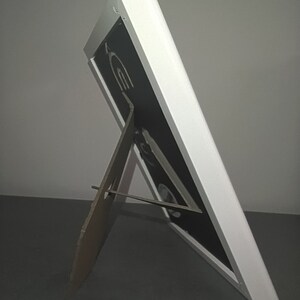 Detailed photo of the backside of a white frame, standing on a table. The backside is black. The frame can be used standing or hanging on the wall.