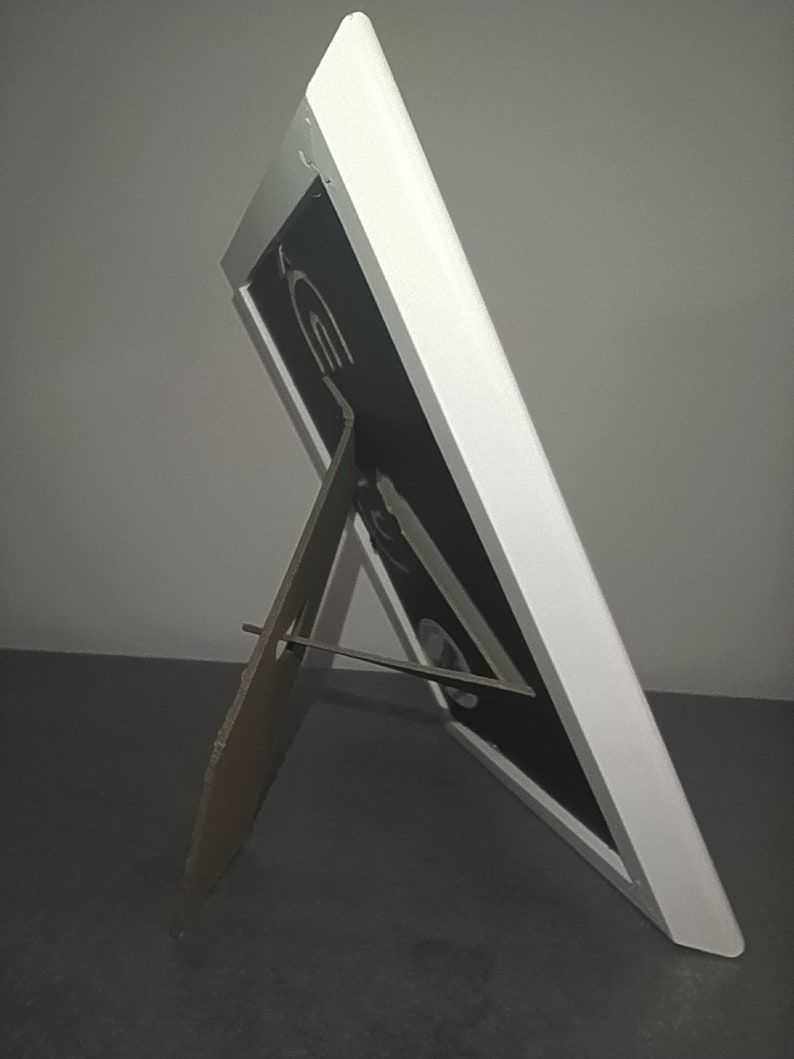 Detailed photo of the backside of a white frame, standing on a table. The backside is black. The frame can be used standing or hanging on the wall.