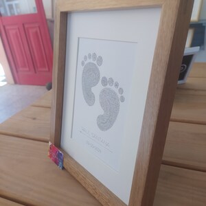 Op de afbeelding: Een ingelijst afbeelding van babyvoetjes in zwart met blauw. De afbeelding is op een witte achtergrond met de naam "Raul Santana" en de datum "09-03-2024" in zwarte inkt.