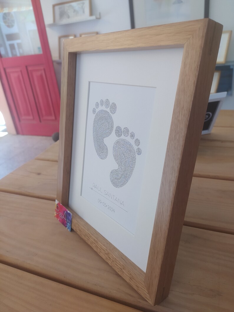 Op de afbeelding: Een ingelijst afbeelding van babyvoetjes in zwart met blauw. De afbeelding is op een witte achtergrond met de naam "Raul Santana" en de datum "09-03-2024" in zwarte inkt.