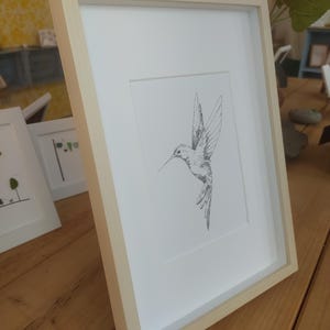 May include: A framed black and white line drawing of a hummingbird in flight. The frame is a light wood color.
