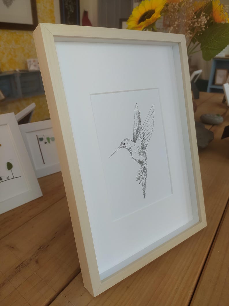 May include: A framed black and white line drawing of a hummingbird in flight. The frame is a light wood color.