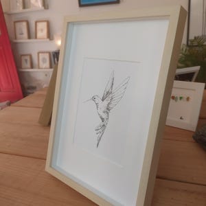 May include: A framed black and white line drawing of a hummingbird in flight. The frame is a light wood color.