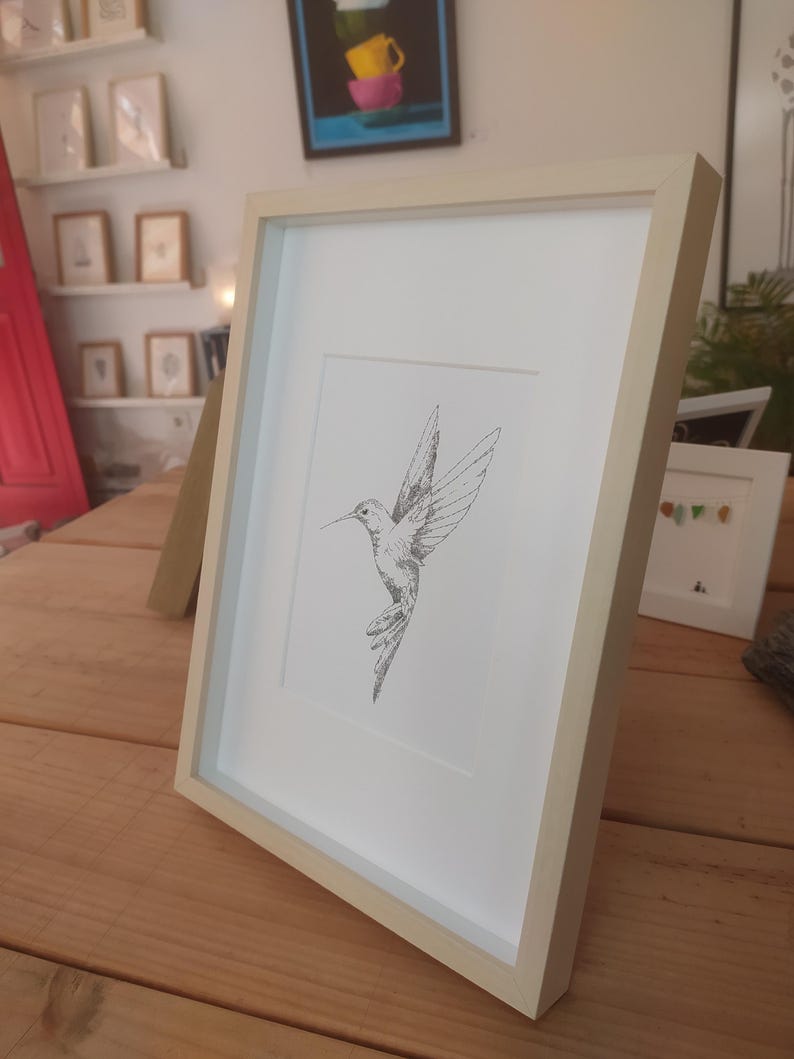 May include: A framed black and white line drawing of a hummingbird in flight. The frame is a light wood color.