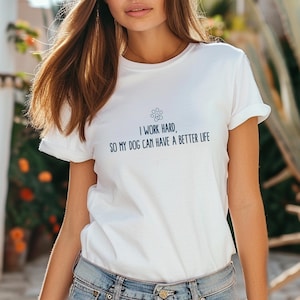 Könnte beinhalten: Weißes T-Shirt mit umgekrempelten Ärmeln und dem marineblauen Text "I WORK HARD, SO MY DOG CAN HAVE A BETTER LIFE". Ein Pfotenabdruck-Grafik befindet sich über dem Text. Das Shirt wird mit einer blauen Jeans kombiniert.