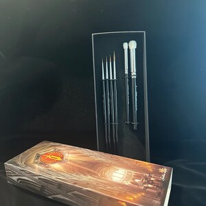 May include: A set of makeup brushes in a black box, with a matching box in the foreground. The brushes have silver handles and black bristles. The box in the foreground has a detailed illustration of a cathedral.