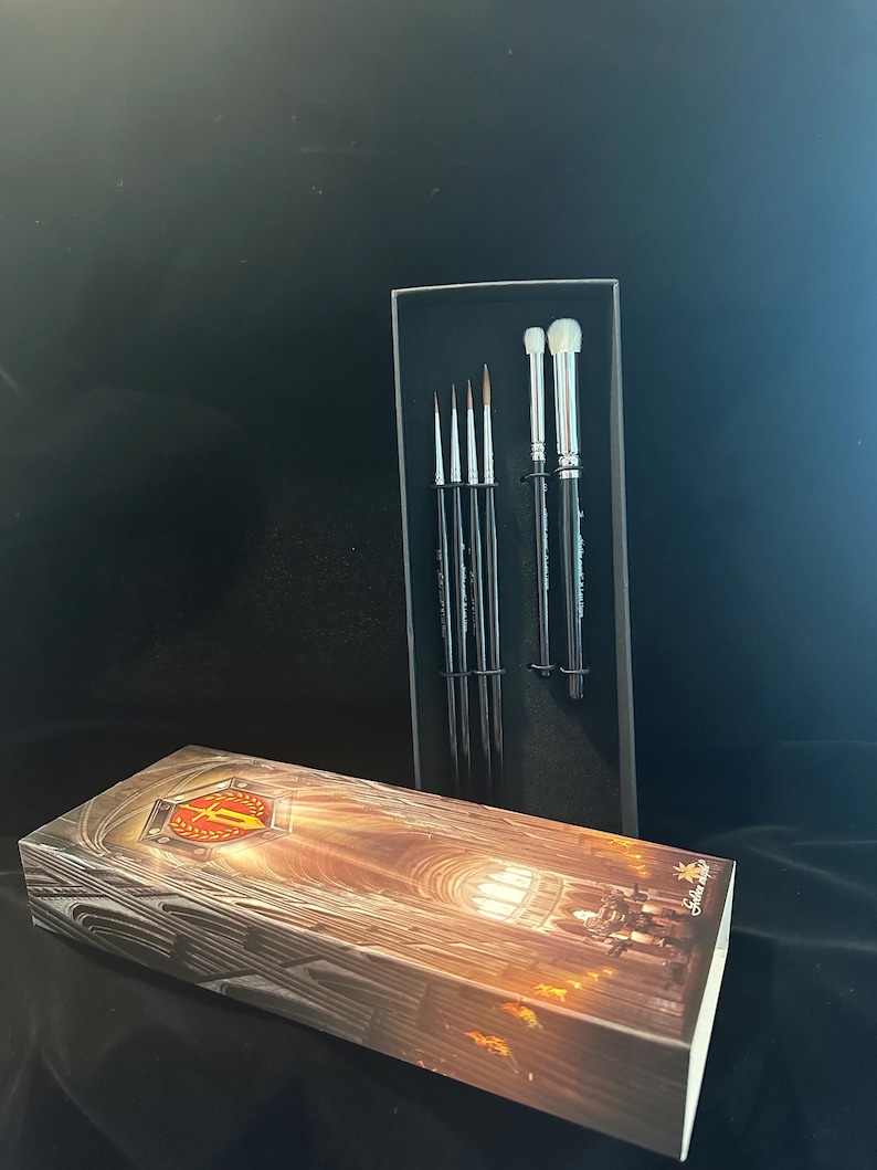 May include: A set of makeup brushes in a black box, with a matching box in the foreground. The brushes have silver handles and black bristles. The box in the foreground has a detailed illustration of a cathedral.