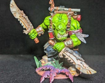 Puede incluir: Una figura miniatura de orco de piel verde que sostiene dos grandes hojas de metal dentadas. El personaje lleva una armadura de cuero marrón con un emblema de calavera, una mochila y se apoya sobre una base decorada con pequeñas plantas verdes y moradas.
