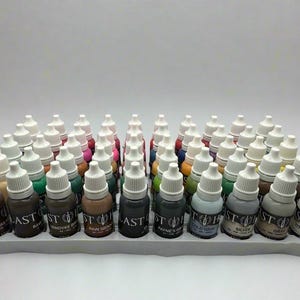 May include: A collection of small paint bottles with white caps, arranged in rows on a gray tray. The bottles display various colors, including brown, green, and gray. Each bottle has a label with the word "LAST" and other text.