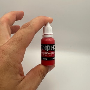 May include: A small, red bottle of "COCHINEAL RED" ink with a white dropper top. The bottle has black text and a logo. The bottle is held in a hand against a white background. The bottle contains 15ml of ink.