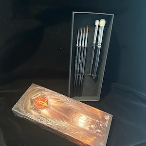 May include: A set of makeup brushes in a black box, with a matching box on the side. The brushes have black handles and silver ferrules. The box on the side has a gold and red emblem.
