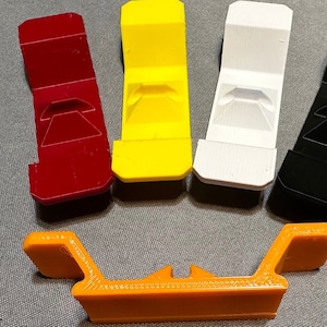 May include: Five 3D printed plastic phone stands in different colors: red, yellow, white, black, and orange. The stands are designed to hold a phone upright.