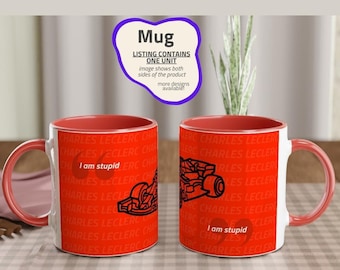 May include: A white ceramic mug with a red rim. The mug has a black and white illustration of a race car and the text "I am stupid" printed on it. The mug is surrounded by a red and white pattern of the text "Charles Leclerc".