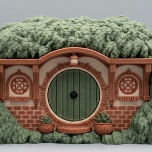 May include: A miniature earth-sheltered dwelling featuring a round green door, a brick facade, and two circular windows. The structure is surrounded by a textured, green, grassy mound and includes two small potted plants flanking the entrance.