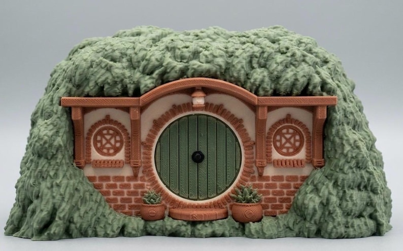 May include: A miniature earth-sheltered dwelling featuring a round green door, a brick facade, and two circular windows. The structure is surrounded by a textured, green, grassy mound and includes two small potted plants flanking the entrance.