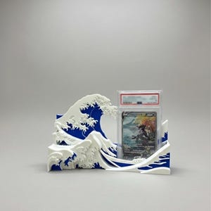 May include: A blue and white plastic display stand shaped like a stylized ocean wave, designed to hold a single rectangular trading card in a clear protective plastic slab. The stand features a layered design with white foam crests over blue.