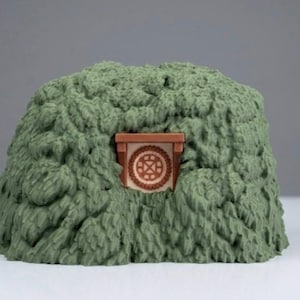 May include: A sculpted green mound with a textured, leafy appearance featuring a small, centered brown door frame. The door is circular with a geometric cross pattern in the center, set against a light-colored background within the foliage.