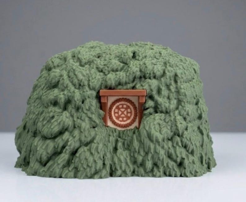 May include: A sculpted green mound with a textured, leafy appearance featuring a small, centered brown door frame. The door is circular with a geometric cross pattern in the center, set against a light-colored background within the foliage.