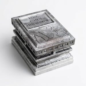 May include: A rectangular silver-toned box with a weathered, stone-like texture. The lid features a raised panel with engraved runic-style characters and a cracked surface pattern. The sides of the box are decorated with a repeating geometric grid.