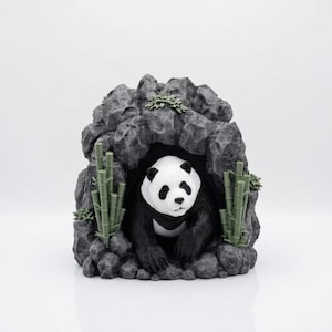 May include: A three-dimensional sculpture of a black and white panda bear emerging from a dark, textured gray rock cave. The scene is decorated with small, light green bamboo stalks on both sides of the cave entrance and tiny green foliage.