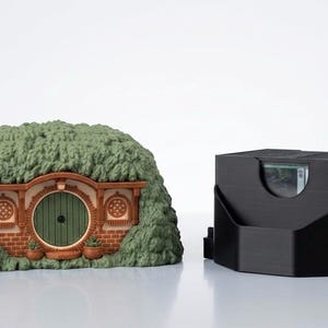 May include: A miniature earth-covered dwelling with a round green door, brick facade, and two circular windows, positioned next to a black rectangular card storage box with a cutout opening that reveals a glimpse of a playing card inside.