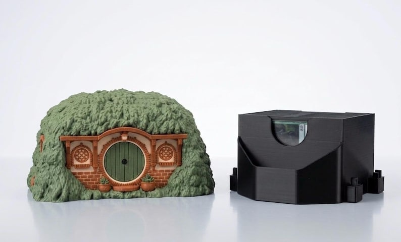 May include: A miniature earth-covered dwelling with a round green door, brick facade, and two circular windows, positioned next to a black rectangular card storage box with a cutout opening that reveals a glimpse of a playing card inside.