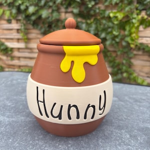 May include: A brown honey pot with a matching lid, featuring a bright yellow drip design on the upper section and a cream-coloured band around the centre. The word Hunny is printed in black, stylised lettering on the cream-coloured band.