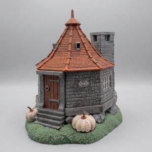 May include: A miniature stone cottage with a conical orange tiled roof and a tall gray chimney. The structure features a wooden door with metal hinges, small windows, and a green base decorated with two light-colored pumpkins on the front.