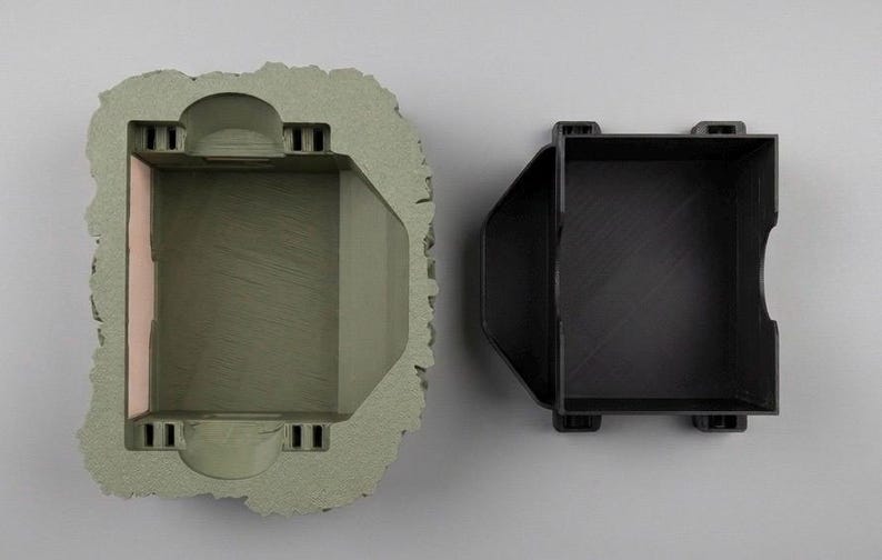 May include: A top-down view of two components: a sage green, textured, rock-like outer shell with a rectangular hollow center, and a separate black, smooth, rectangular insert with a notched side designed to fit within the outer shell.