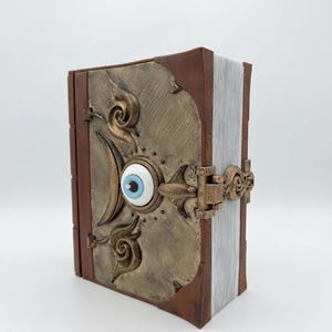 May include: A brown book with a metallic gold-toned cover plate featuring a central blue and white eye ornament. The cover has embossed swirl patterns and a hinged metal clasp. The pages are visible on the side with a light gray textured finish.