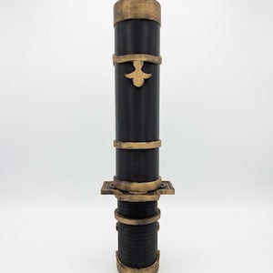 May include: A cylindrical black tube featuring metallic gold-colored bands and decorative accents. The object includes a prominent gold fleur-de-lis emblem on the upper section and a rectangular base plate near the center, with a textured finish.