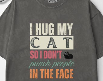 May include: Dark grey t-shirt featuring the text "I HUG MY CAT SO I DON'T PUNCH PEOPLE IN THE FACE" in white, pink, and orange. A black cat graphic is also included. The shirt has a crew neck and short sleeves.