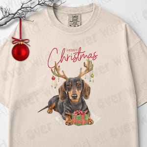 May include: Ivory-colored t-shirt with a dachshund wearing antlers and holding a gift. The words "Merry Christmas" are written above the dog. The shirt has a crew neck and short sleeves. The brand logo "All Over Wild" is in the bottom left corner.